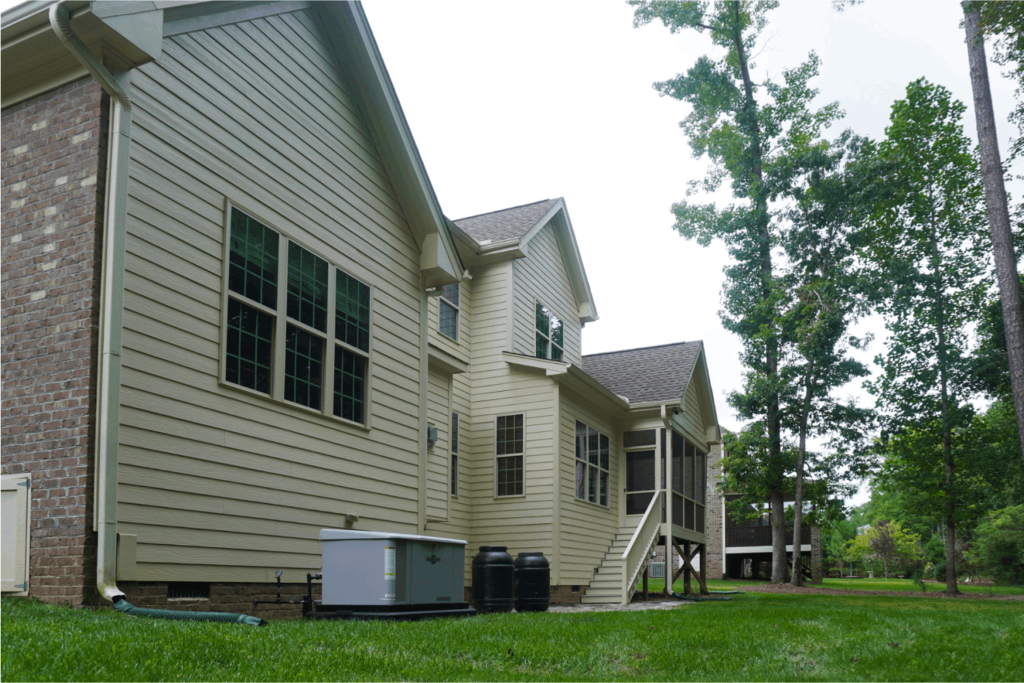 View of the back of a tan home with green grass and tall trees in backyard.
