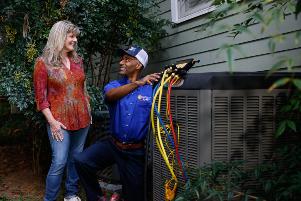 Technician speaking with homeowner while kneeling next to an outdoor HVAC unit next to a home with green siding. Tall bush behind them.