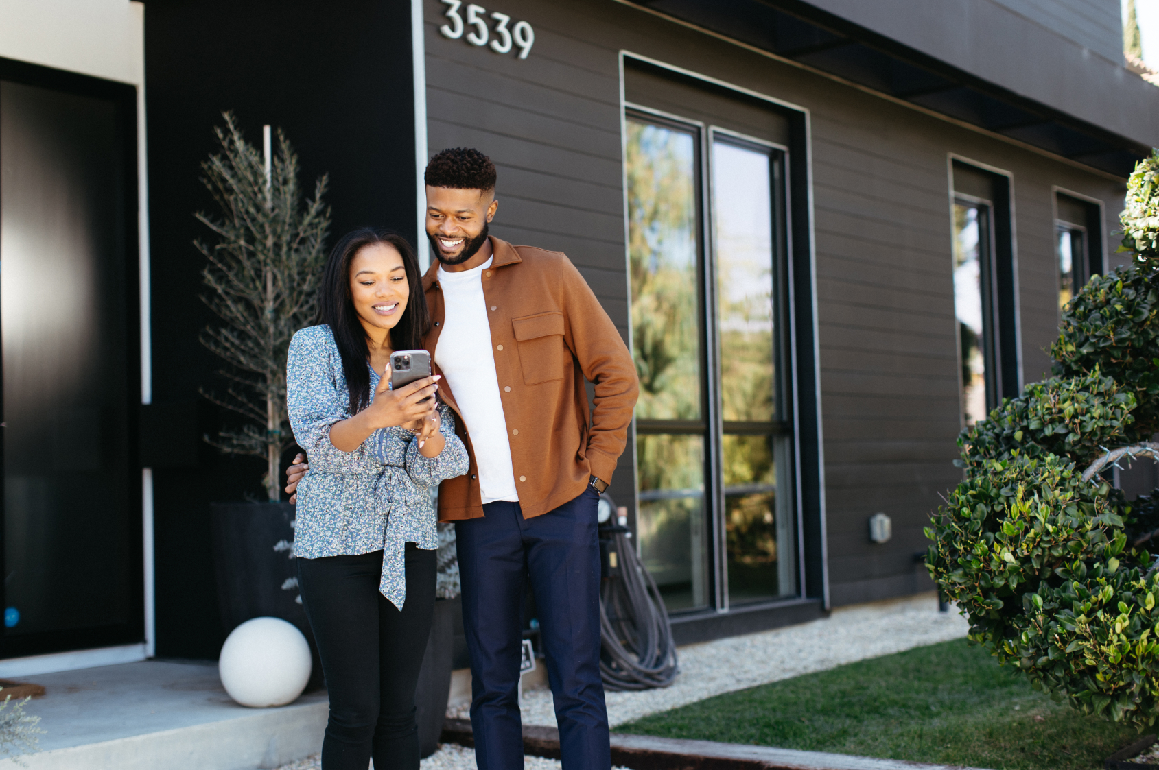 Homeowners smiling and looking at a phone together, standing in front of a home with black siding.